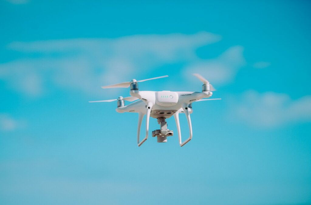 A white camera drone hovering in a blue sky with scattered clouds