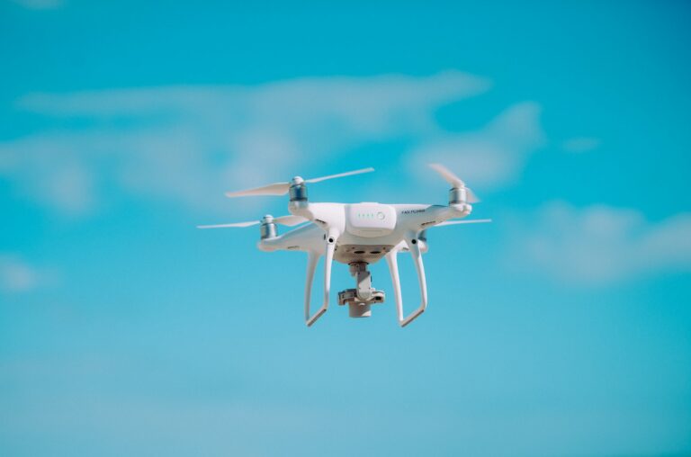 A white camera drone hovering in a blue sky with scattered clouds